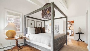 Una cama con dosel sobre cuatro postes, con estructura de madera rústica, una mesita de noche tejida con una lámpara y vistas a la habitación contigua.