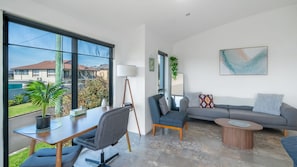 A modern living room with a sofa, a coffee table, a desk with a chair, and large windows offering a view of the outside.
