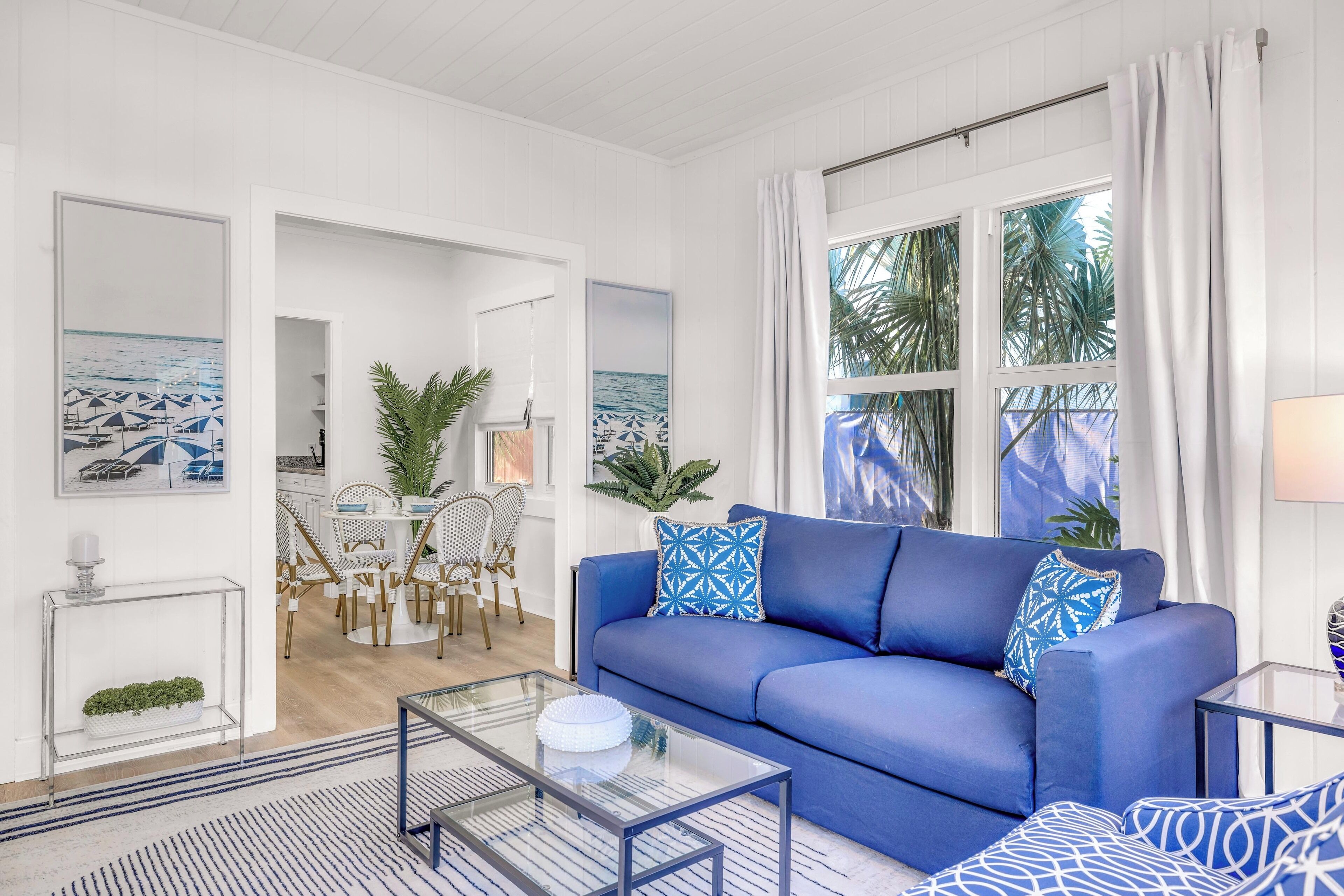 A modern living room with a blue sofa, a glass coffee table, and a dining area with wooden chairs and a white table.