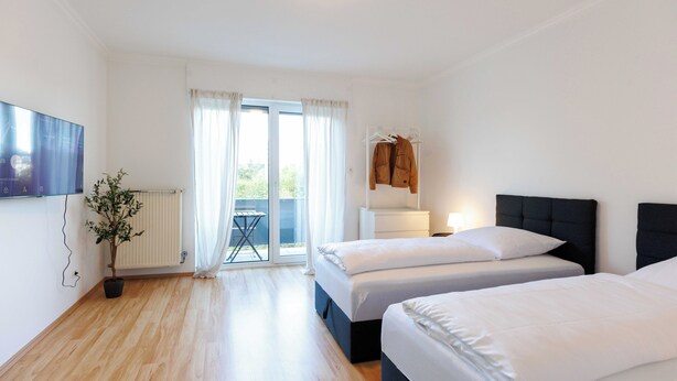 A hotel room with two beds, a TV, a potted plant, and a view of the outdoors through a sliding door. - Apartmenthaus - Kaiserslautern (Kaiserslautern)