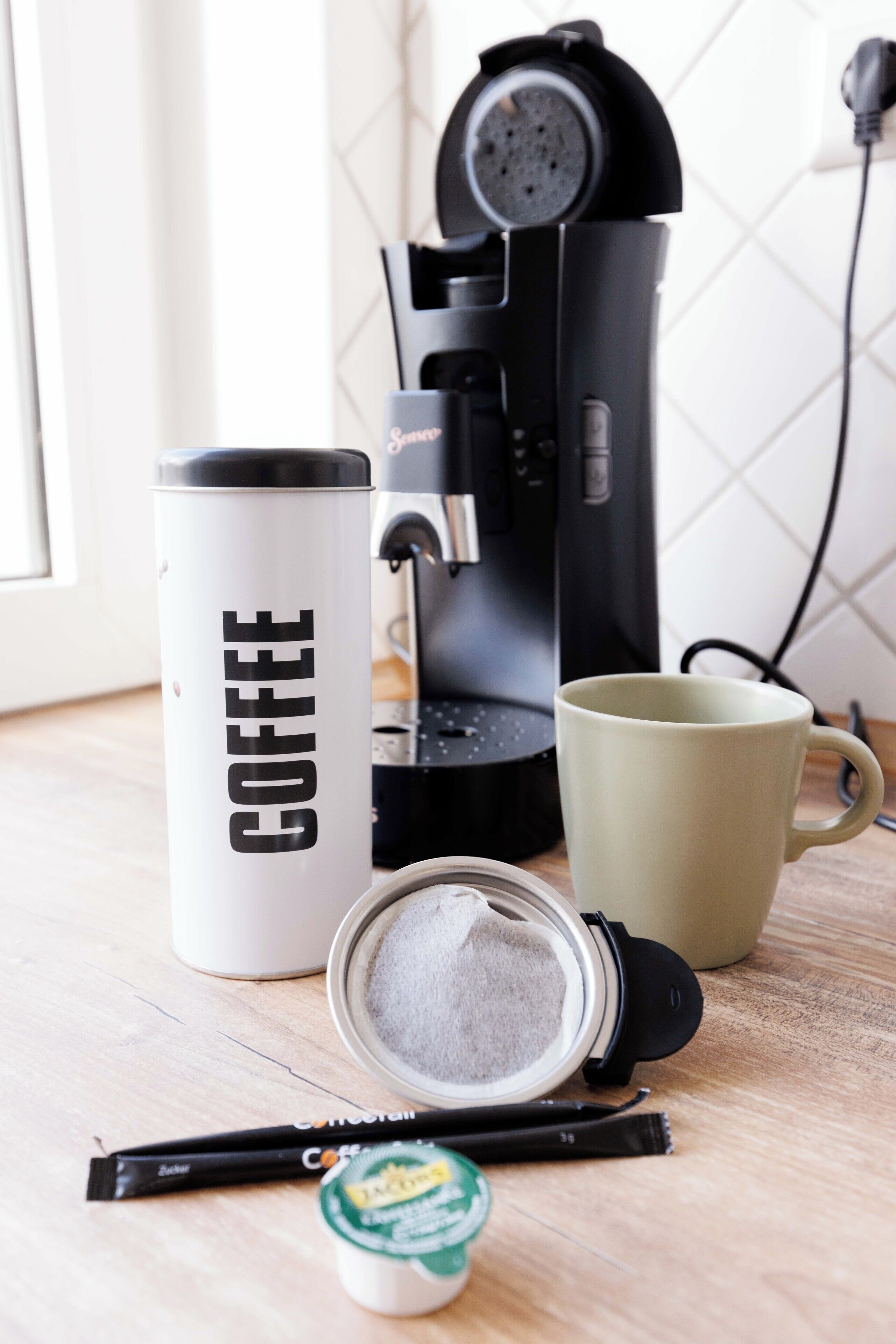 A coffee maker, a mug, a coffee container, and coffee pods on a wooden surface.