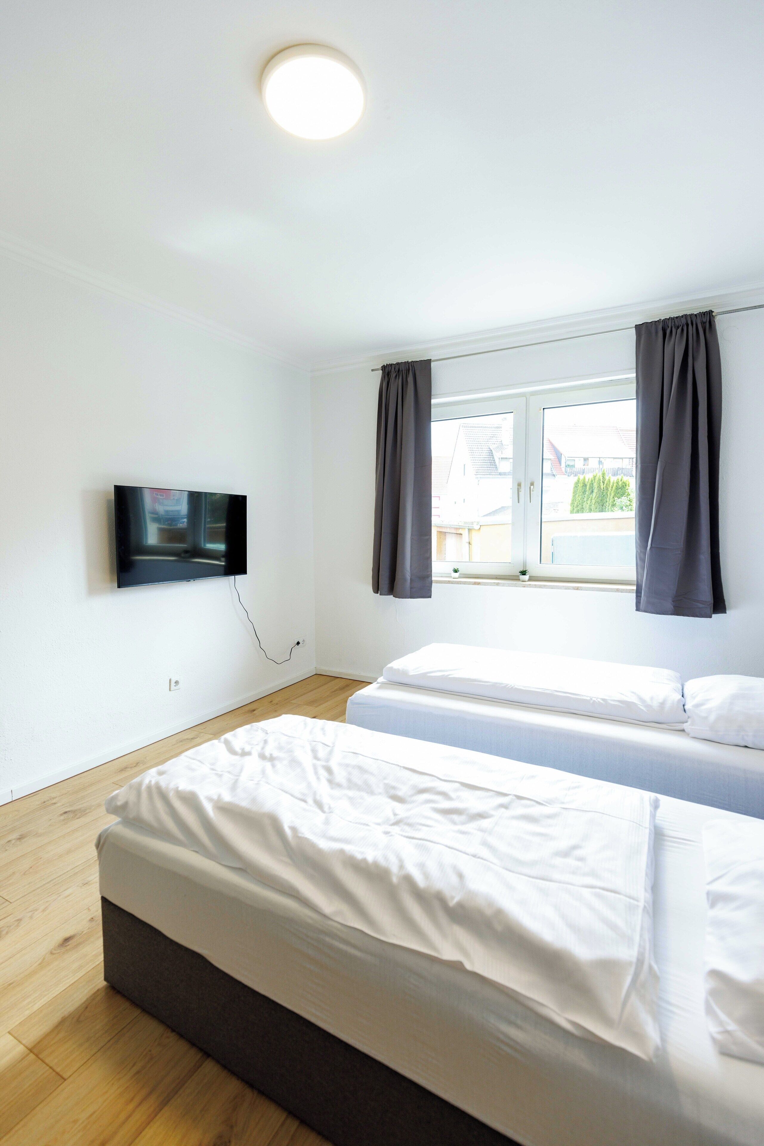 A room with two beds, a TV mounted on the wall, a window with curtains, and a ceiling light.