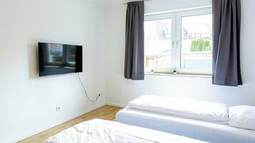 A room with two beds, a TV mounted on the wall, a window with curtains, and a ceiling light.