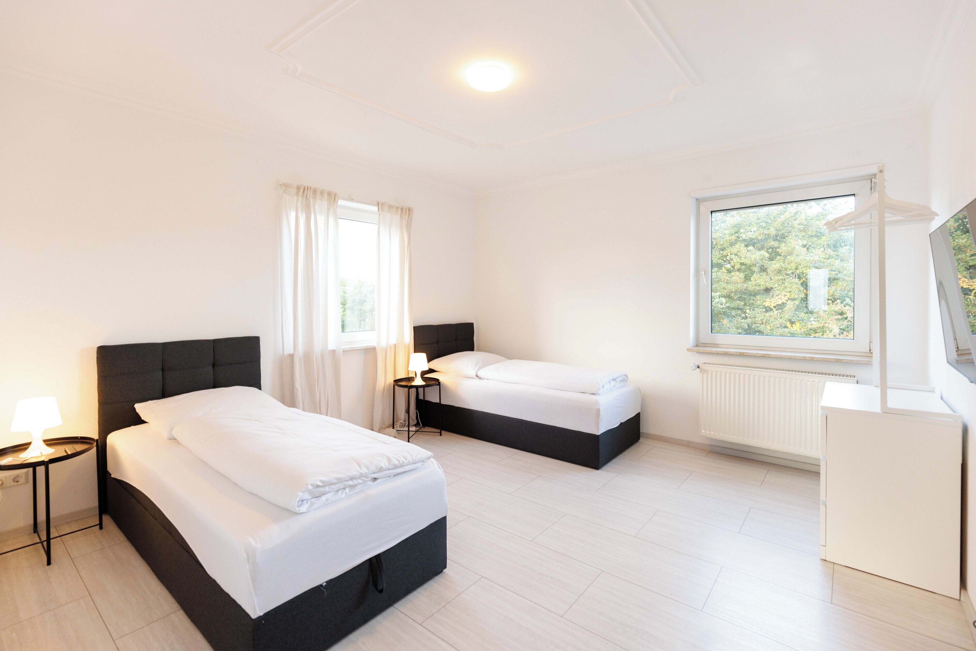 Habitación de hotel con dos camas, una mesita con una lámpara, una ventana con vistas a árboles y un radiador blanco.