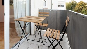A balcony with a wooden table and chairs, a sliding glass door, and a grey wall. - Apartmenthaus - Kaiserslautern (Kaiserslautern)
