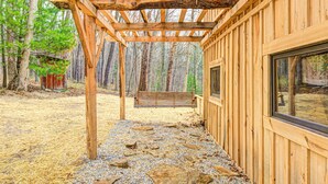 Une cabane en bois avec un patio dallé de pierres, une balançoire en bois et une fenêtre donnant sur la forêt.