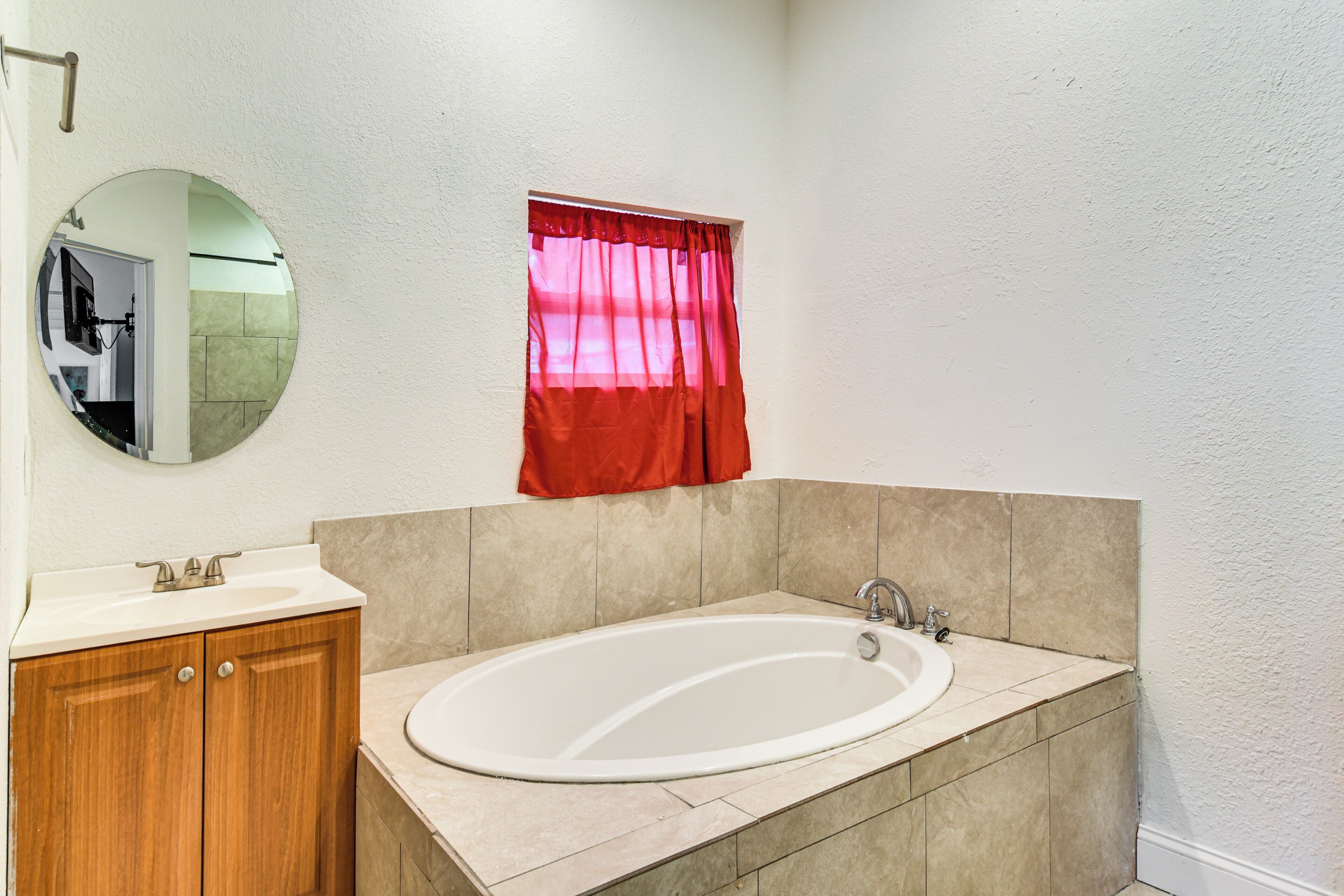 Ein Badezimmer mit einer weißen Badewanne, Holzschränken, einem runden Spiegel und einem Fenster mit roten Vorhängen.
