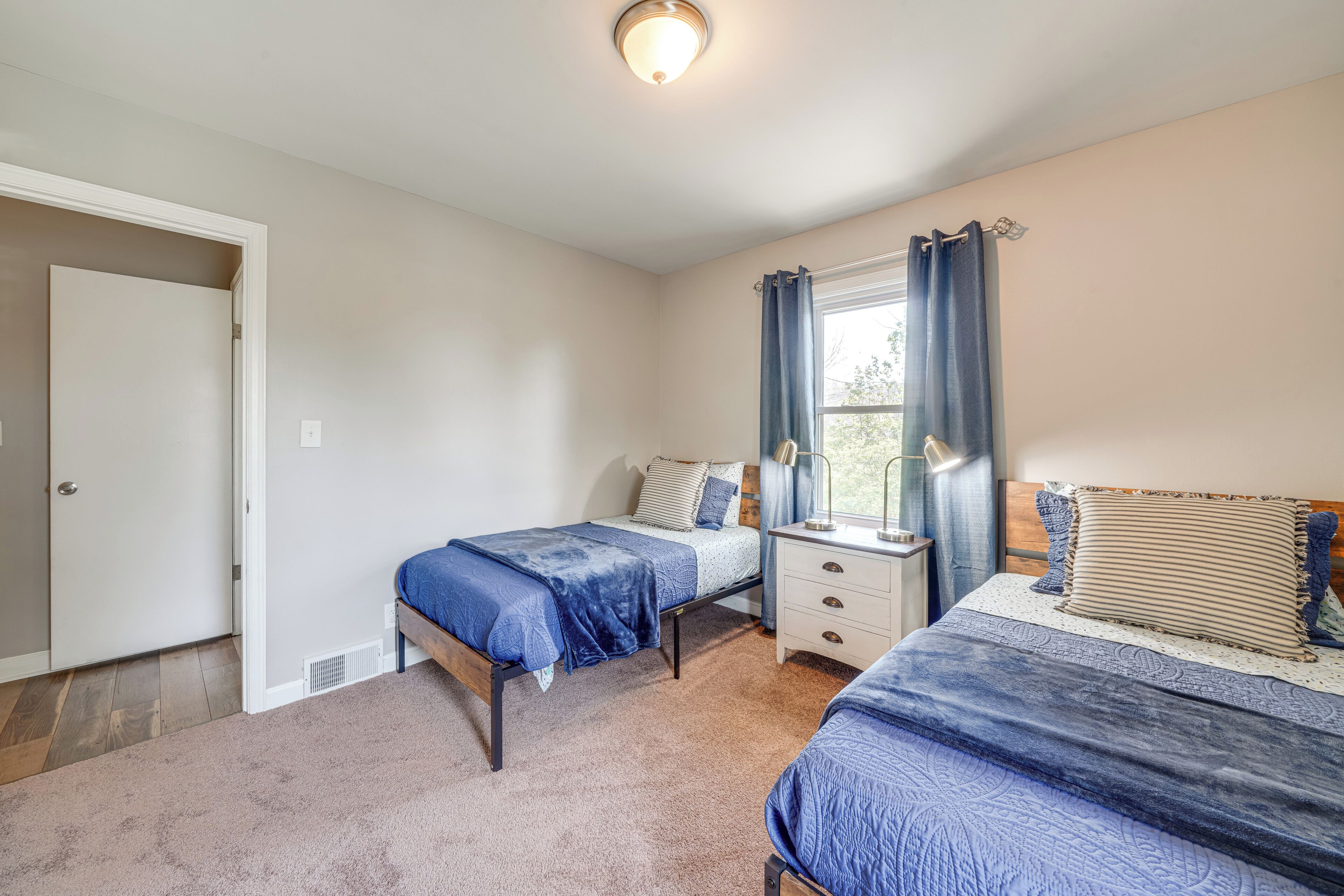 A bedroom with two beds, a nightstand, a window with curtains, and a door.