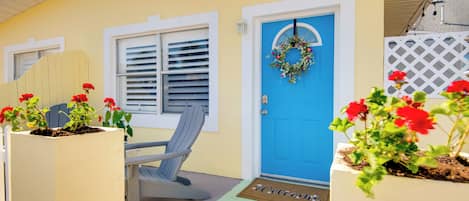 A yellow house with a blue door, a bench, and potted plants with red flowers.