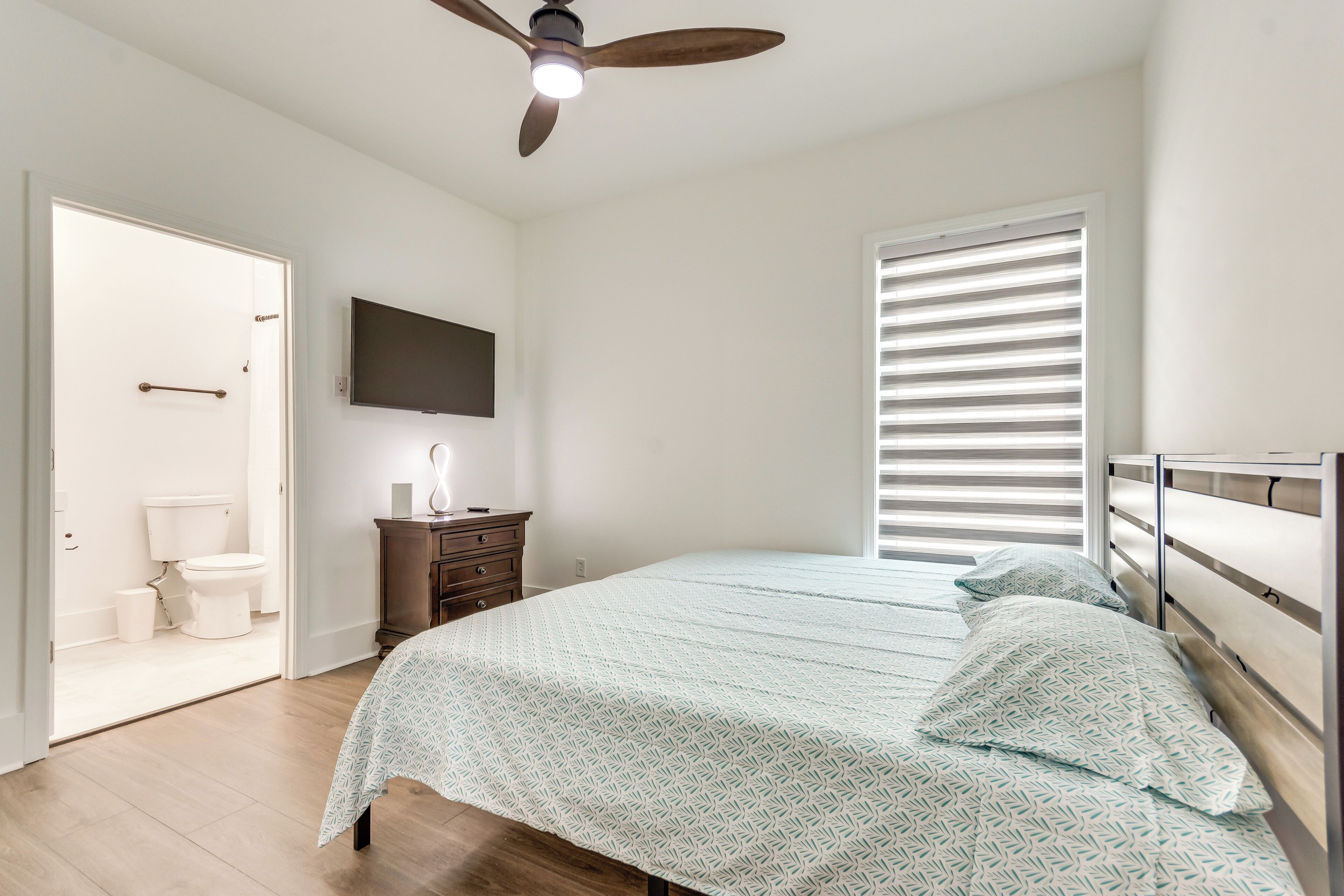 A bedroom with a bed, a ceiling fan, a TV mounted on the wall, and a window with blinds.