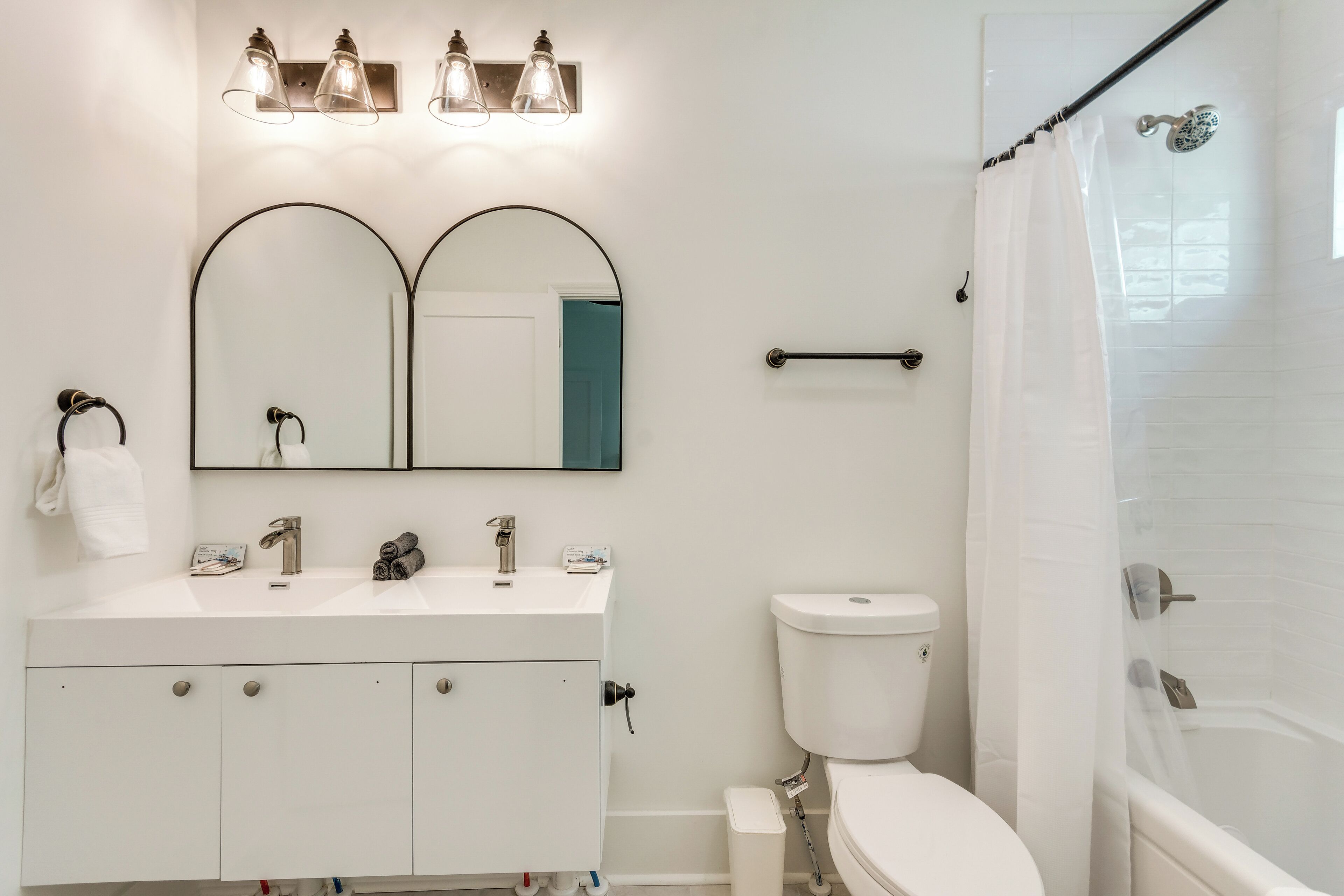 Une salle de bain moderne avec un lavabo blanc, deux miroirs, des toilettes et une douche avec un rideau.