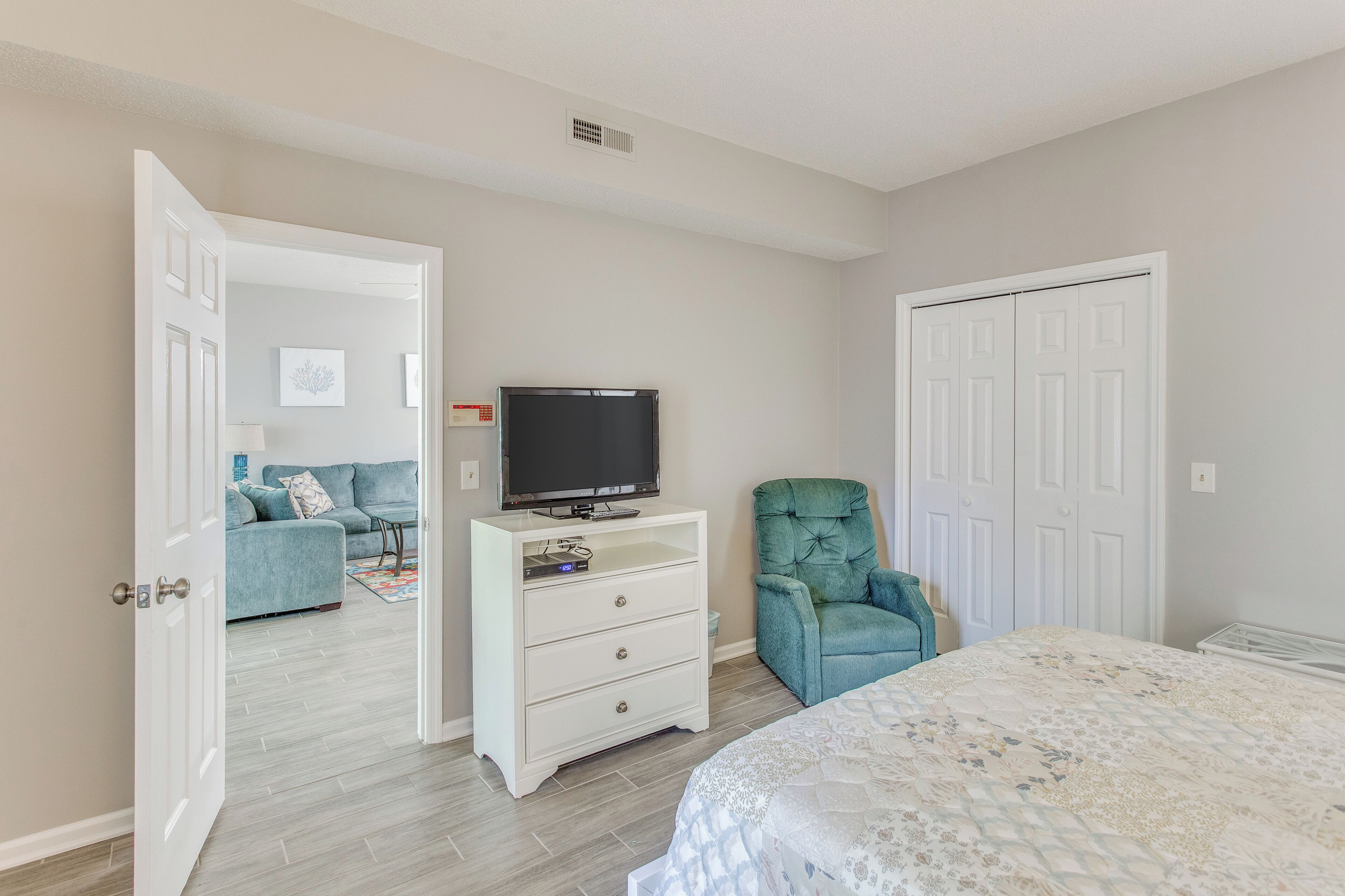 A bedroom with a bed, a dresser, a television, a chair, and a door leading to another room.