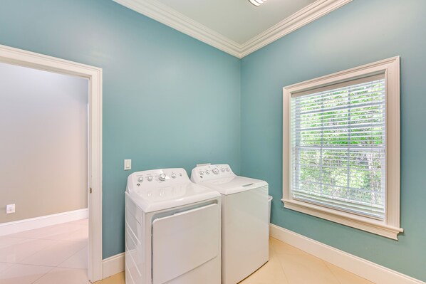 A laundry room with two white front-loading washers and dryers, a window with blinds, and a door leading to another room.
