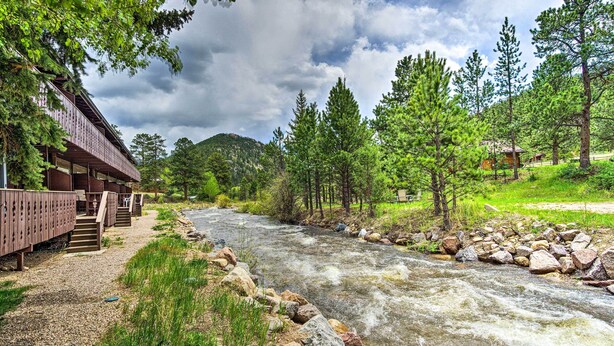 A riverside lodge with a wooden deck and stairs leading to the entrance, surrounded by lush greenery and a flowing river.