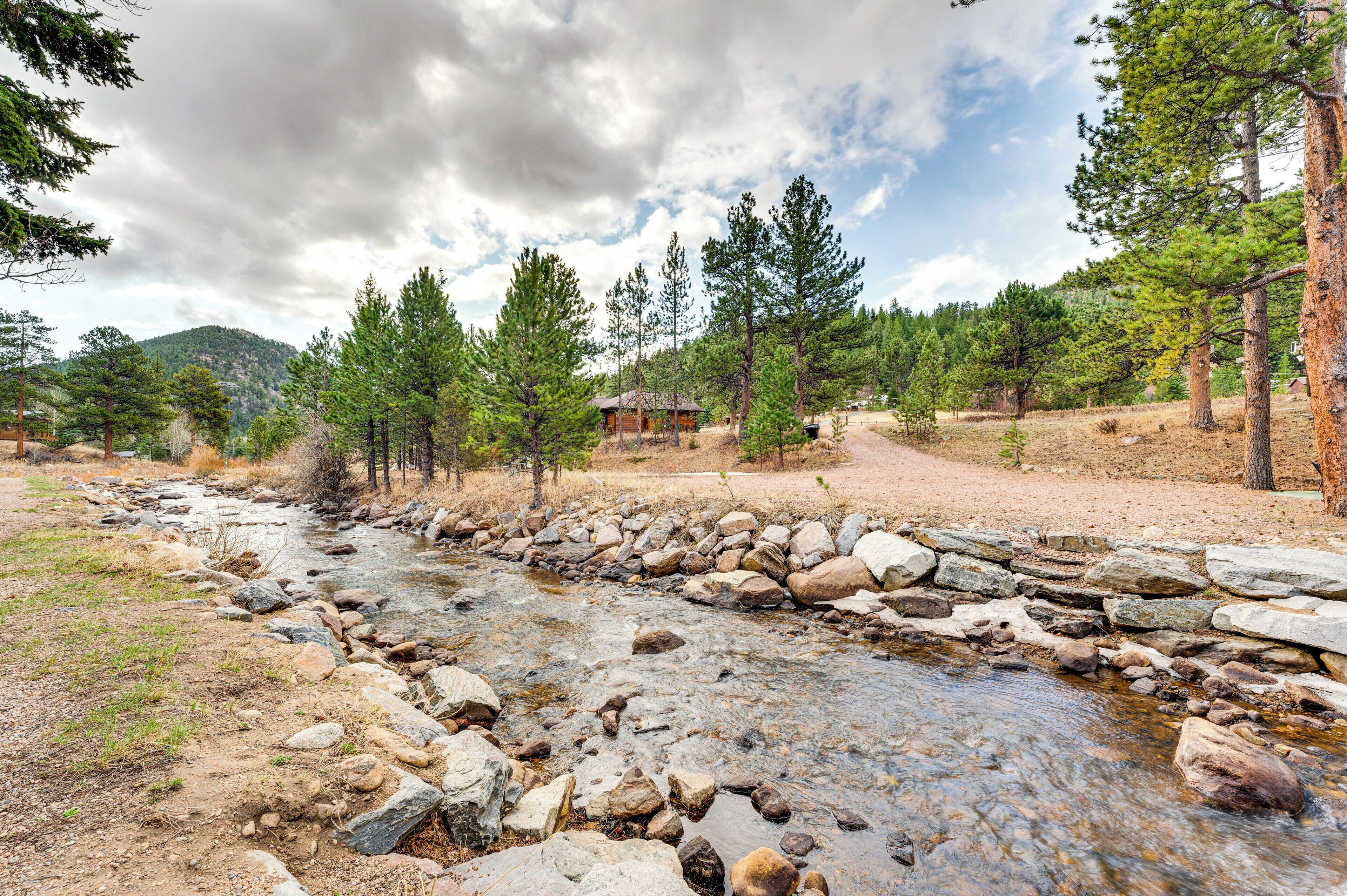 A rustic cabin surrounded by a forest, a rocky stream, and a mountain in the background.