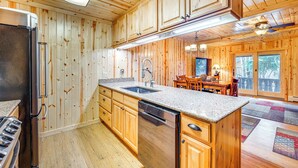 A wooden cabin interior with a kitchen featuring a refrigerator, dishwasher, granite countertop, wooden cabinets, and a dining area with a table and chairs.