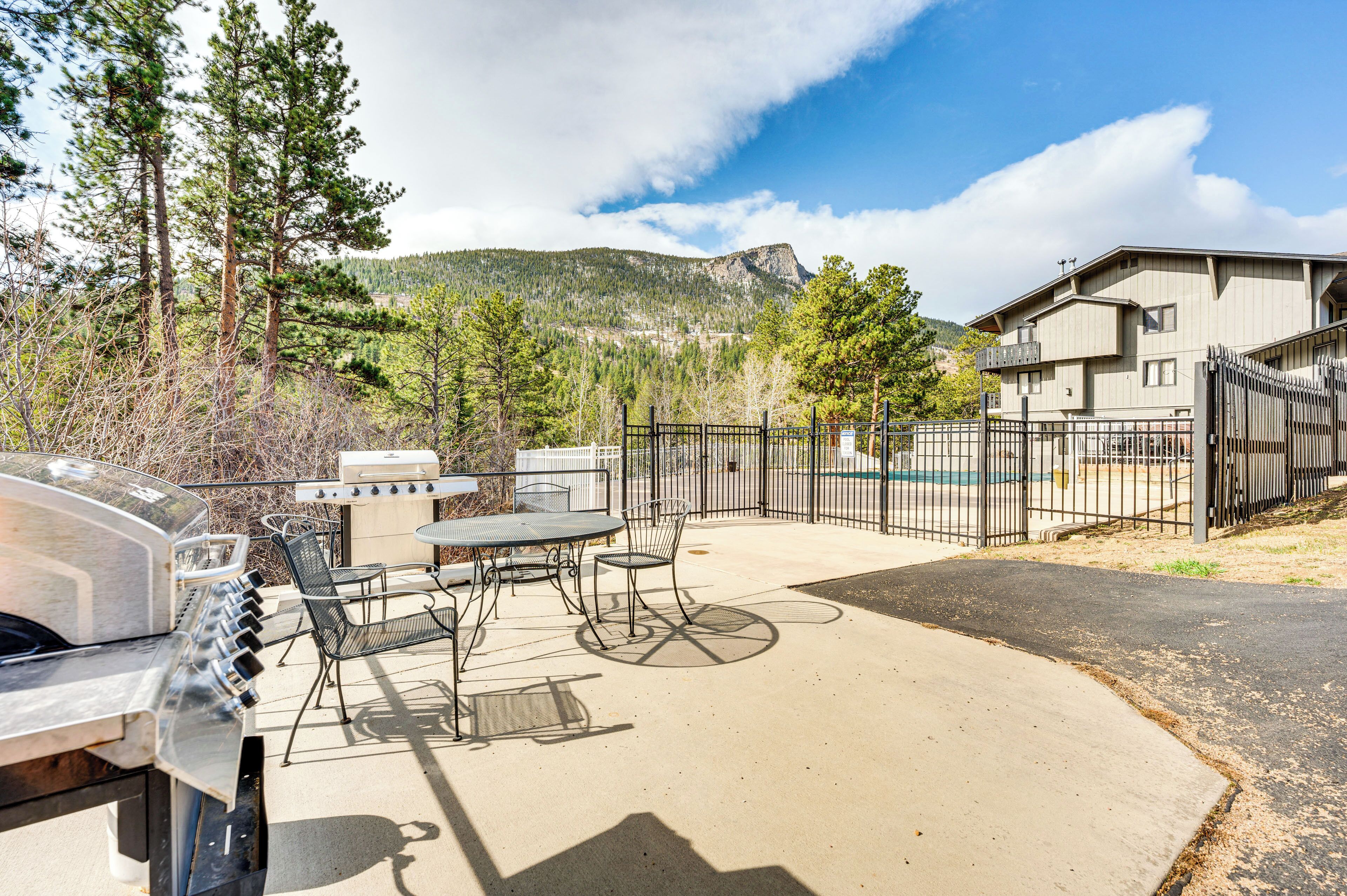 An outdoor patio with a grill, chairs, and a table, surrounded by a fence and a mountain view.