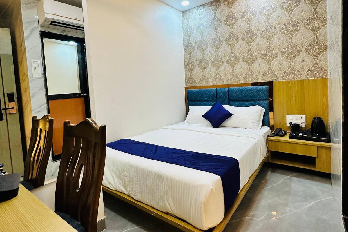 A hotel room with a bed, bedside table, chair, desk, and an air conditioning unit.