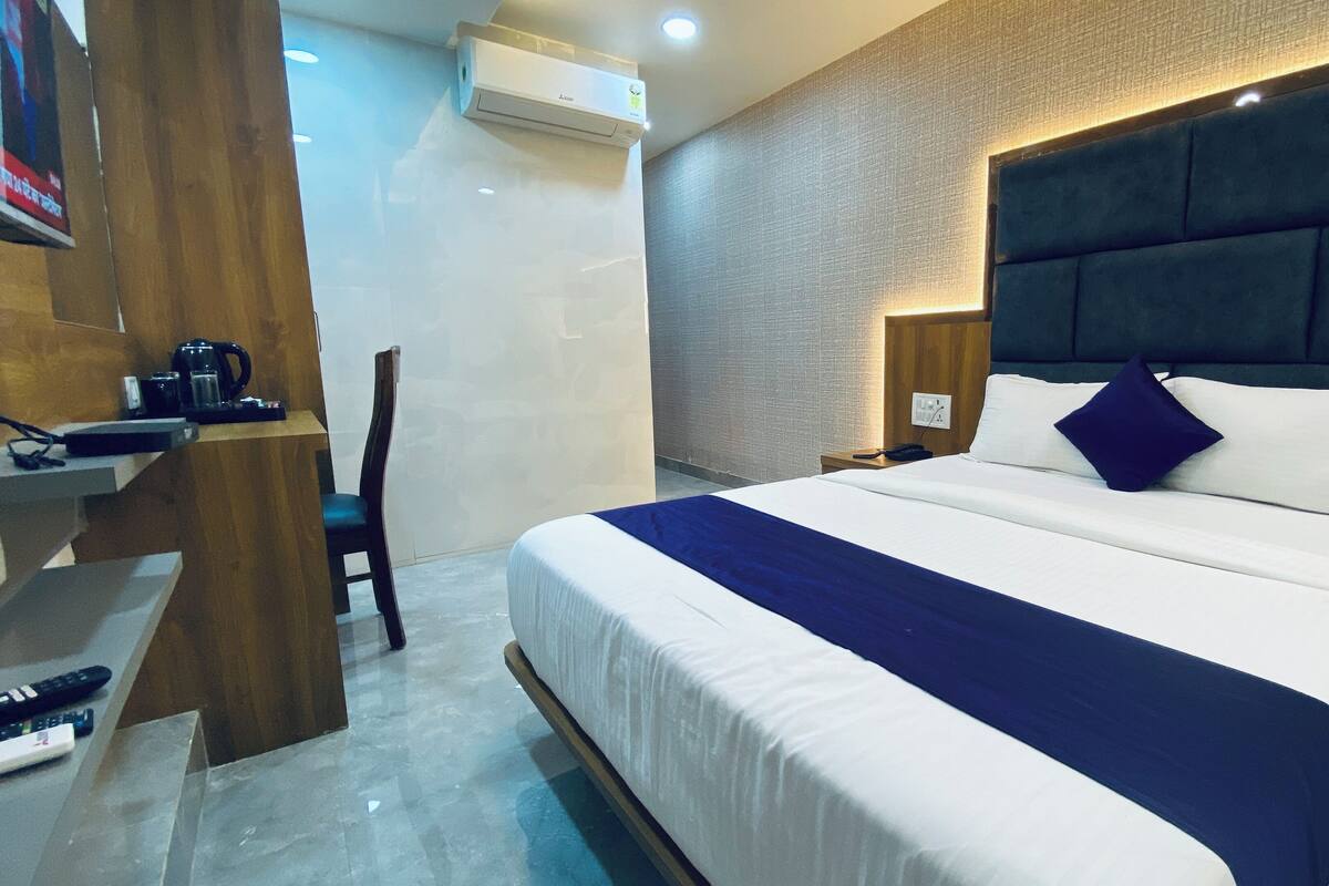 A modern hotel room with a large bed, a wooden nightstand, a desk with a coffee maker, and an air conditioning unit.