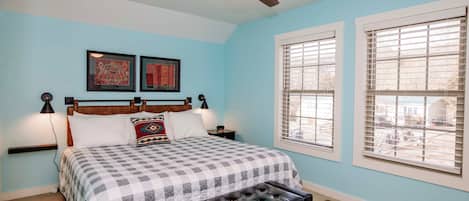 A bedroom with a bed, a ceiling fan, two windows with blinds, and artwork on the wall.