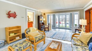 Een ruime woonkamer met een groot raam, een rieten bank, een houten boekenkast en een eethoek met een tafel en stoelen.