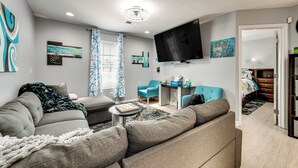 A modern living room with a grey sofa, a small coffee table, and a flat-screen TV mounted on the wall. There is a blue armchair and a desk with a chair in the background. - Near Speedway & Shops: Concord Apt w/ Shared Yard! (Concord)