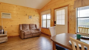 Un acogedor interior de cabaña con suelo de madera, un sofá, una mesa de comedor con sillas y ventanas grandes.