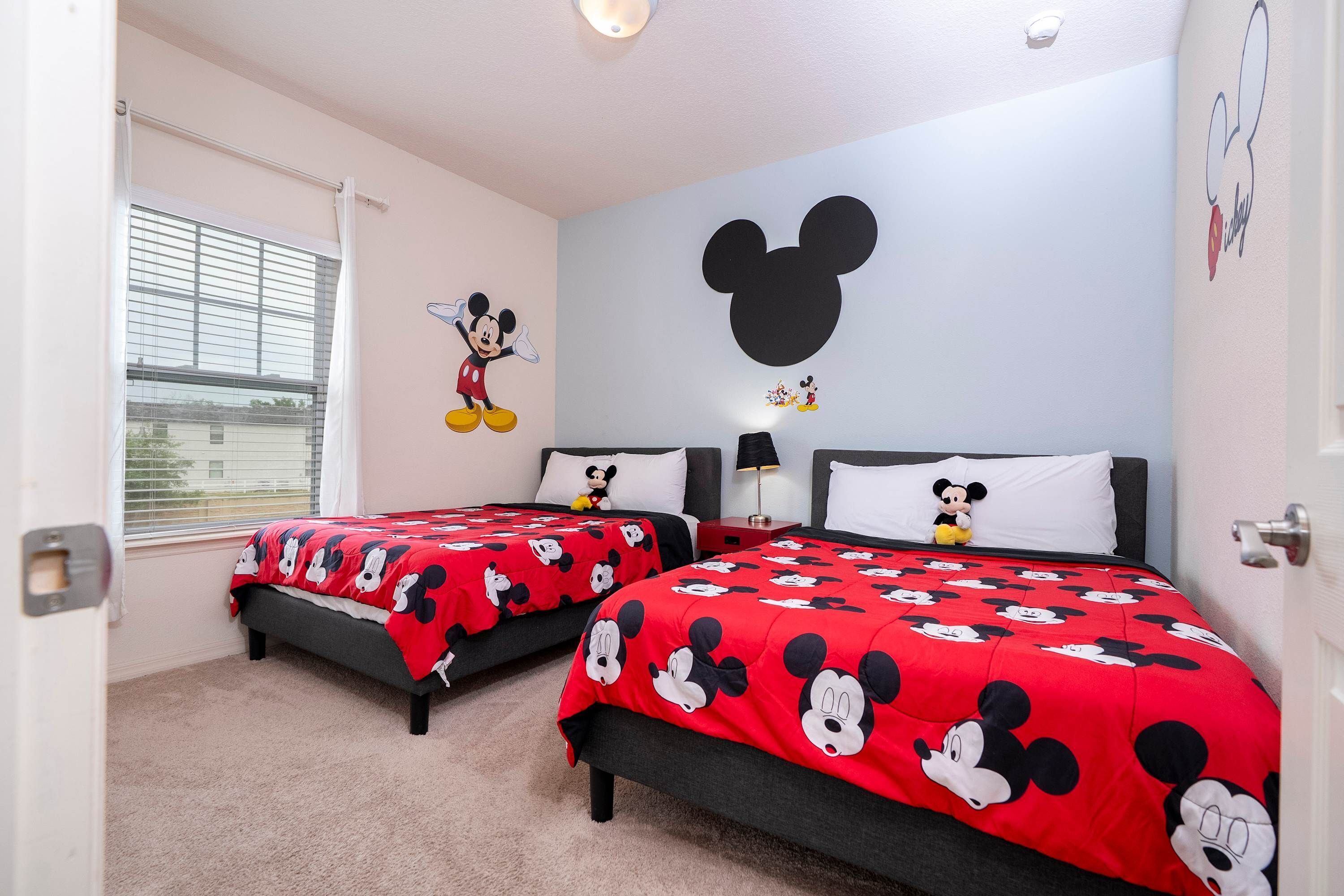 Un dormitorio con dos camas, cada una con ropa de cama temática de Mickey Mouse, un gran adhesivo de pared de Mickey Mouse y una ventana con persianas.