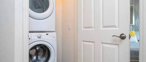 A laundry area with stacked washers and dryers, a white door, and a visible bed with a yellow pillow in the adjacent room.