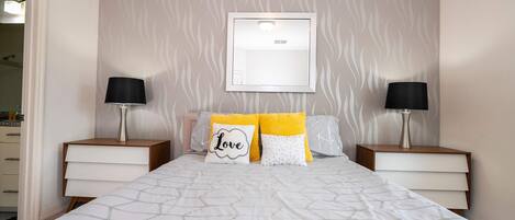Une chambre à coucher bien rangée, avec un lit, deux tables de chevet, une lampe fixée au mur et un tableau encadré au-dessus du lit.