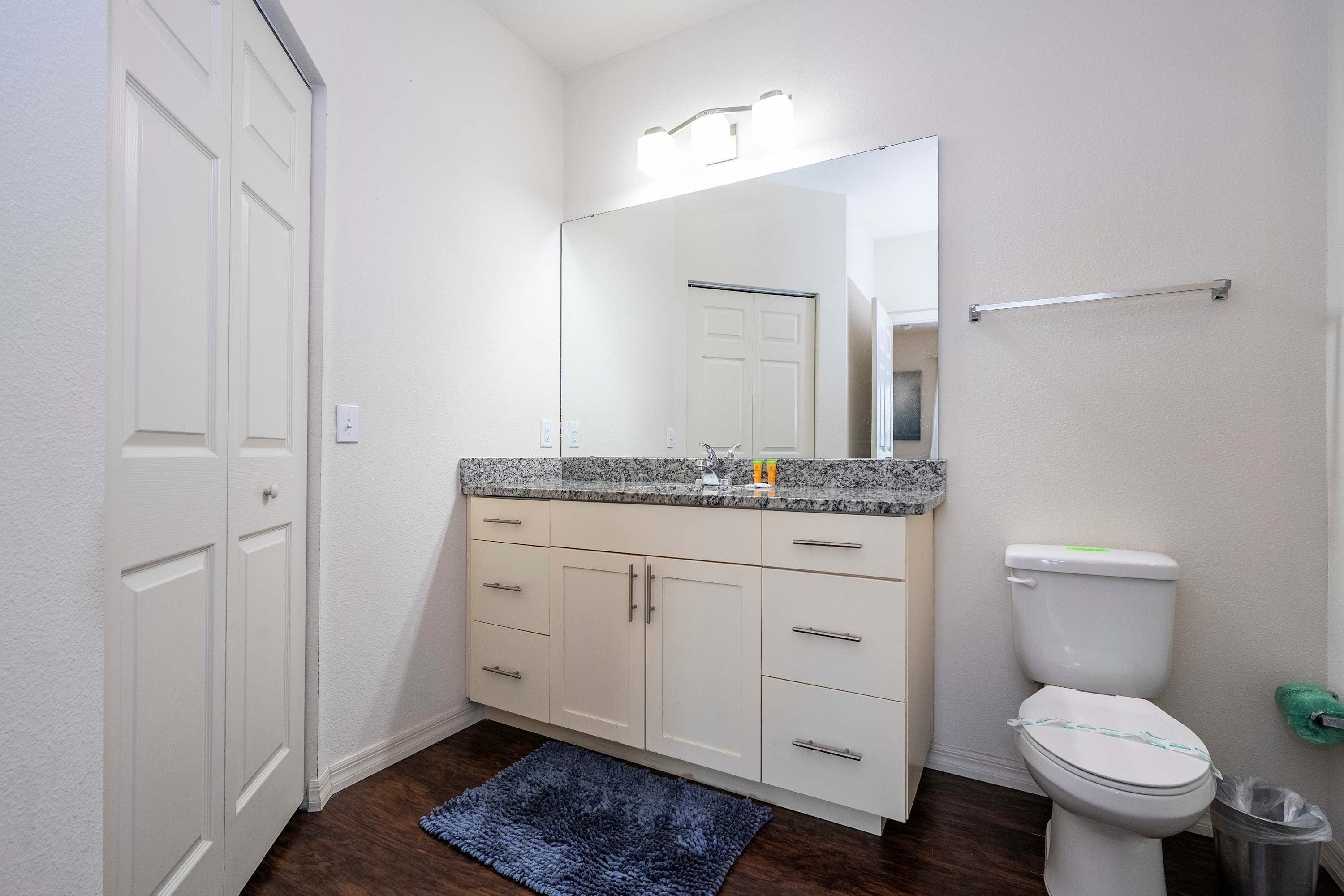 A bathroom with a white toilet, a granite countertop, a mirror, and a blue rug.