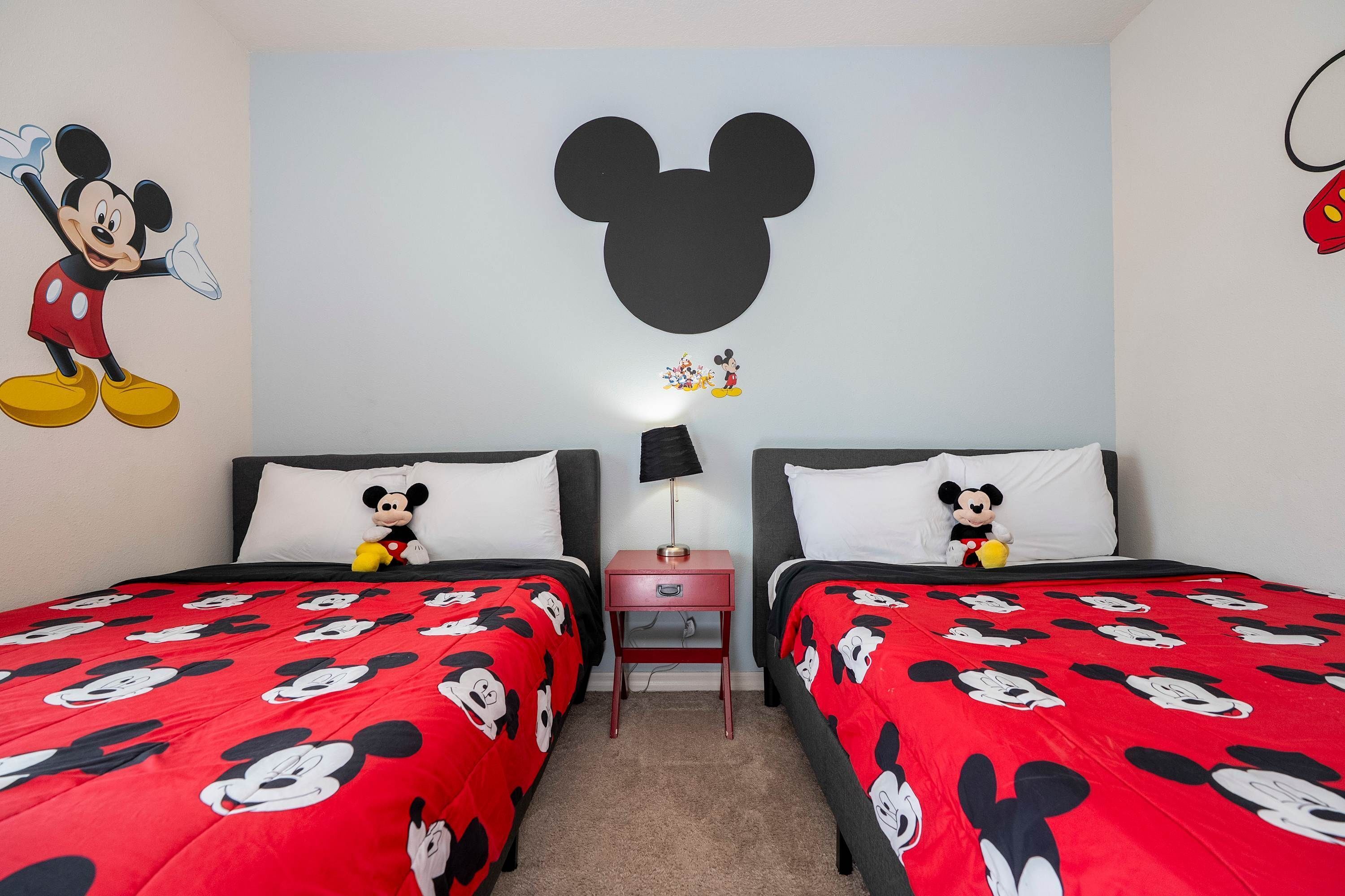 A hotel room with two beds, each with Mickey Mouse themed bedding. There is a large Mickey Mouse head decal on the wall above the beds, and a Mickey Mouse plush toy on each bed.