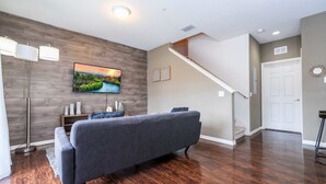 Un salon moderne avec un canapé gris, un plancher en bois, un téléviseur fixé au mur et un escalier menant à un autre niveau.