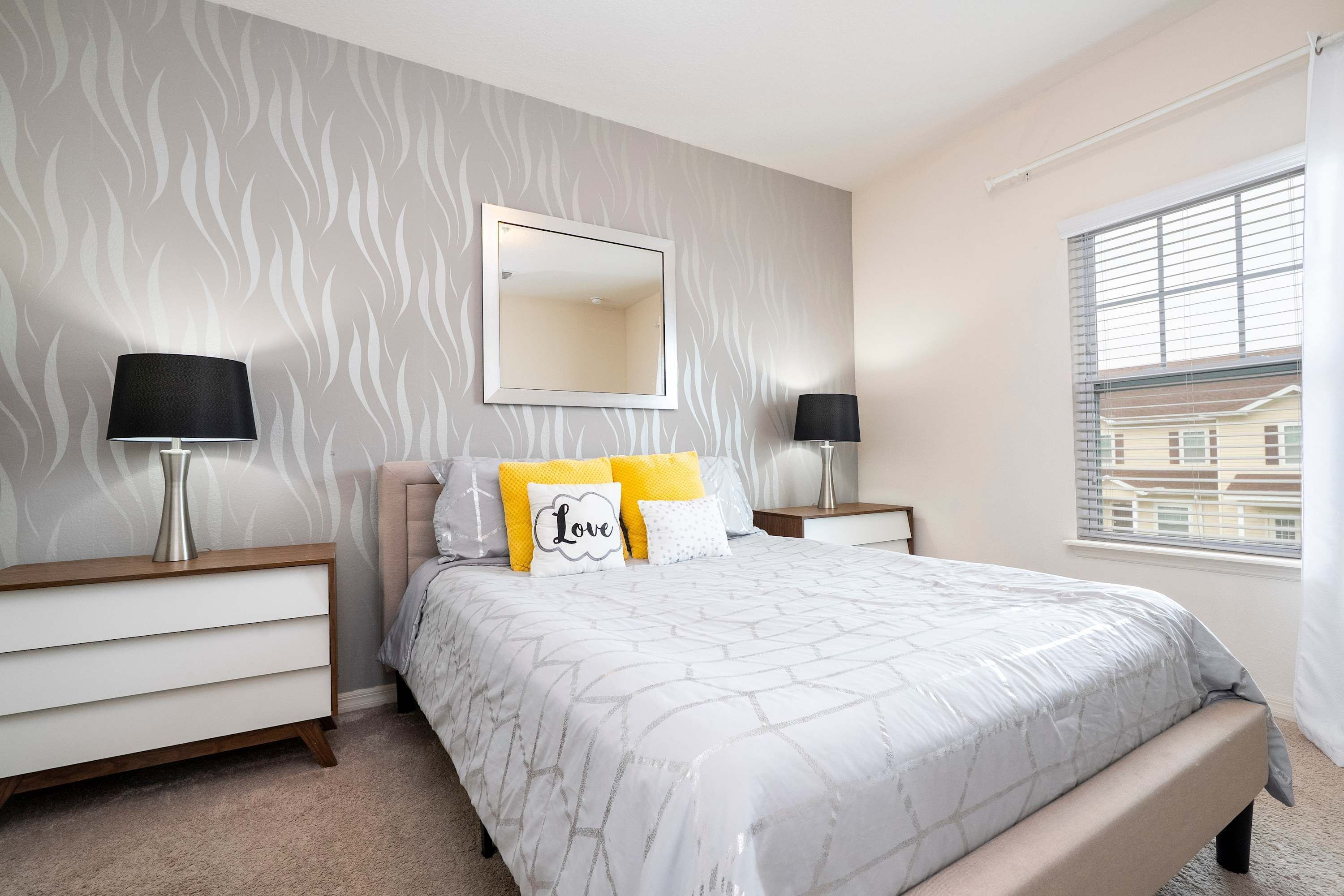 A bedroom with a bed, two bedside tables, a mirror, and a window with blinds.