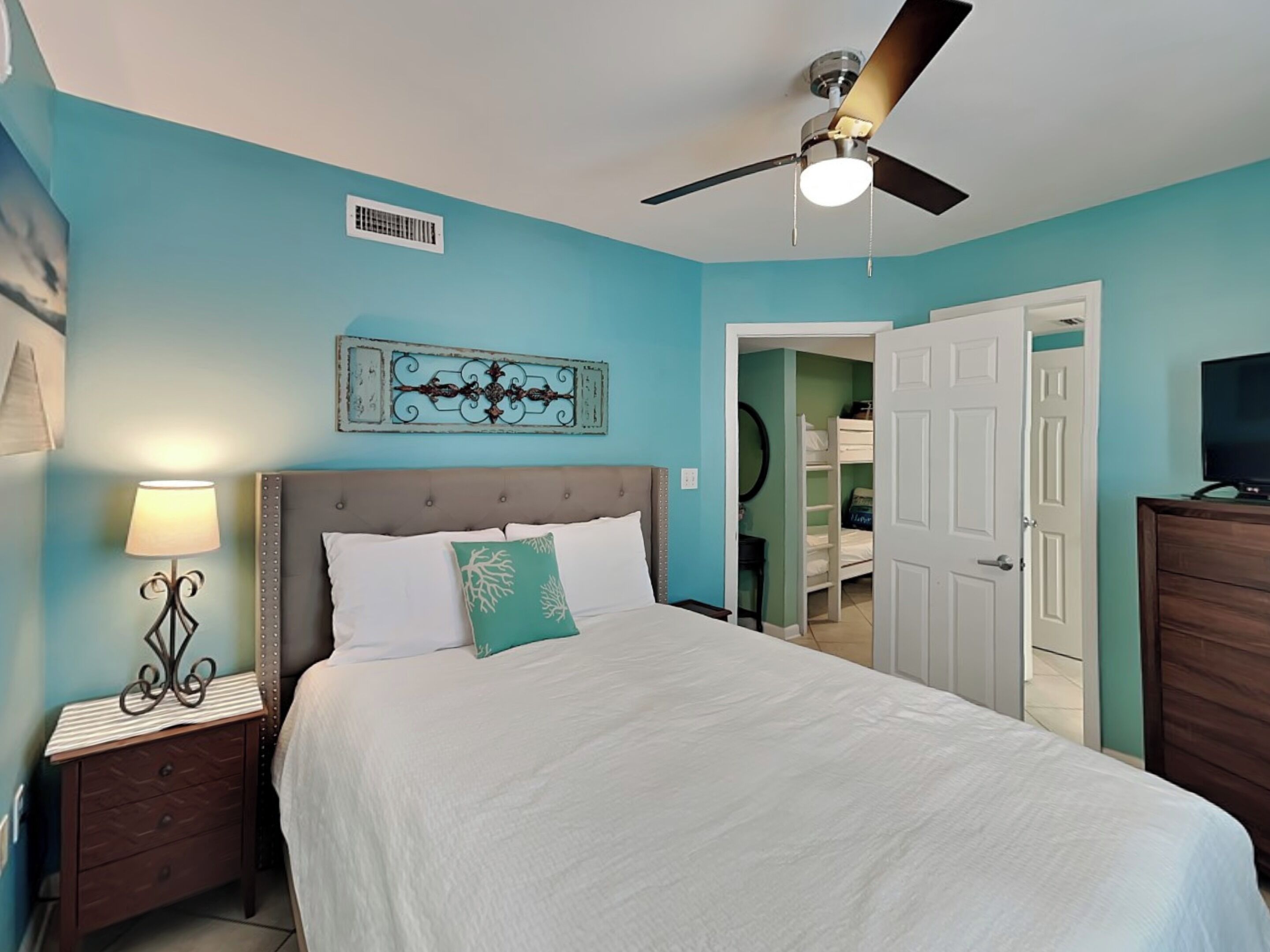 A bedroom with a bed, a nightstand, a lamp, a ceiling fan, a decorative wall piece, a door, and a dresser.