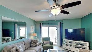 Un salon aux murs de couleur turquoise, doté d’un ventilateur de plafond, d’un canapé, d’un fauteuil, d’une table basse, d’un téléviseur et d’un meuble de rangement blanc pour les appareils multimédias.