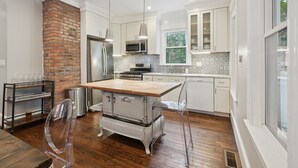 Une cuisine avec des meubles blancs, un mur en briques, des appareils électroménagers en acier inoxydable et une table en bois.