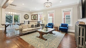 Un vaste salon comprenant une table basse en bois, un canapé et deux fauteuils bleus. On y trouve également un coin repas avec une table et des chaises en verre, ainsi qu’un lustre suspendu au plafond.