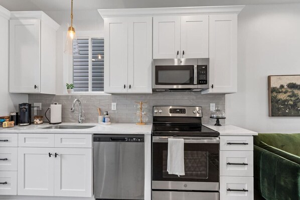 Cocina moderna con armarios blancos, microondas de acero inoxidable, horno, lavavajillas y una silla tapizada de color verde.