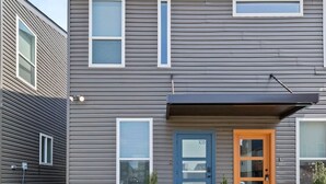 Un immeuble résidentiel moderne à deux étages, avec une façade grise, une porte bleue et un petit jardin devant, agrémenté de plantes.