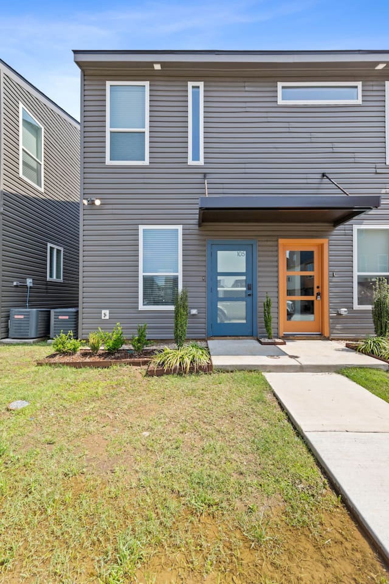 Un immeuble résidentiel moderne à deux étages, avec une façade grise, une porte bleue et un petit jardin devant, agrémenté de plantes.