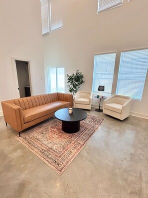 Un salon spacieux avec un canapé en cuir marron, deux fauteuils blancs, une table basse ronde et un tapis à motifs.