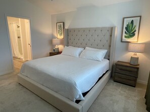 A bedroom with a large bed, a nightstand with a lamp, and a framed plant on the wall.