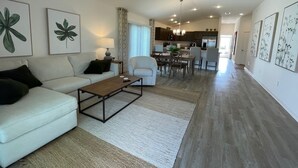 Ein modernes Wohnzimmer mit einer Couch, einem Couchtisch und Kunstwerken. Im Hintergrund ist ein Essbereich mit Stühlen und einem Tisch zu sehen.