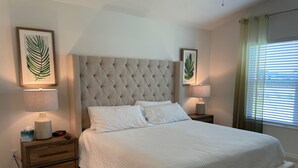 Ein ordentlich eingerichtetes Schlafzimmer mit einem gepolsterten Kopfteil, einem großen Bett mit weißer Bettwäsche, zwei Nachttischen mit Lampen und gerahmten Pflanzenmotiven an den Wänden.