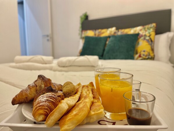 Bandeja de desayuno con cruasanes, pan y jugo sobre una cama con almohadas y cabecera.