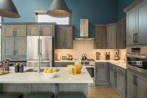 Une cuisine avec des meubles gris, un réfrigérateur en acier inoxydable, un grand îlot central orné d’un saladier rempli de citrons et une carafe de thé glacé.