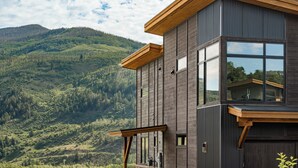 Ein modernes Gebäude mit großen Fenstern, einer Holzterrasse und einer Bergkulisse im Hintergrund.