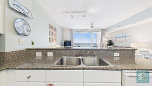 A modern kitchen with granite countertops, stainless steel sinks, and a view of the ocean through large windows. - Seabreeze 721-Gulf/Pool Views-Sleeps 8 (Panama City Beach)