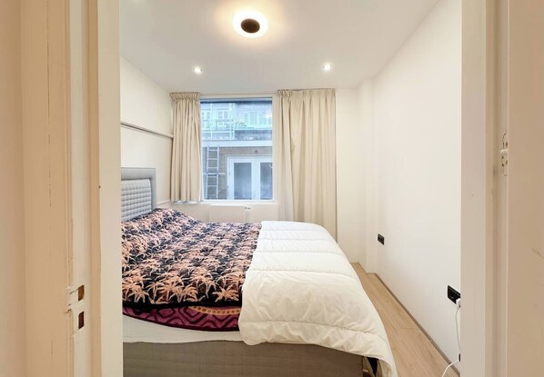 A bedroom with a bed, a window with curtains, a radiator, and a view of a building outside.